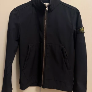 Jacka Stone Island - Säljer en snygg svart jacka från Stone Island i mycket bra skick. Jackan har en dragkedja framtill och två praktiska fickor. Den ikoniska loggan sitter på ärmen. Perfekt för höst och vår! Storlek 12 år/ 156. 