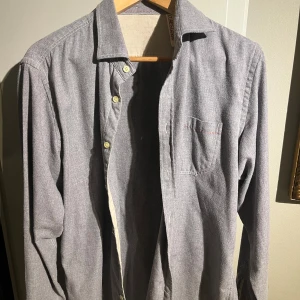 Grå långärmad skjorta från Claes Göran - Säljer en stilren grå långärmad skjorta i bra skick. Skjortan har en klassisk krage och knappar framtill. Perfekt för både vardag och festliga tillfällen. Passar bra till jeans eller chinos för en avslappnad look.