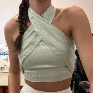 Grön omlott topp - Snygg grön omlott topp med blommigt mönster. Den har en unik omlott-design som ger en stilren look. Perfekt för sommardagar eller en festlig kväll. Toppen är ärmlös och har en tight passform som framhäver figuren.