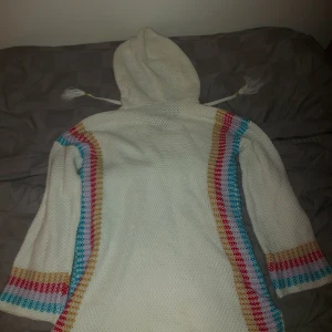 Vit stickad hoodie med färgglada ränder - Säljer en mysig vit stickad hoodie med färgglada ränder i rött, blått och beige. Den har en stor ficka fram och en luva med snören. Perfekt för kyliga dagar när du vill vara både bekväm och stilren. Passar bra till både vardag och avslappnade tillfällen.