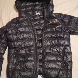 Svart pufferjacka från Emporio Armani - Säljer denna billihare då den har ett hål vid luvan på höger axel, men har suttit en tejp bit där som jag även målat svart, men det går lätt att sy ihop .