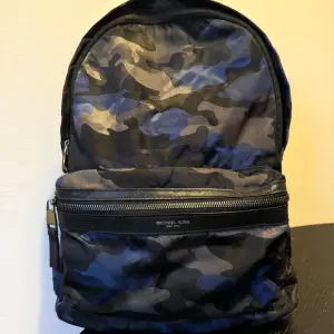 Säljer en snygg ryggsäck från Michael Kors i ett coolt blått och svart camouflage-mönster. Den har en praktisk dragkedja framtill och justerbara axelremmar. Perfekt för vardagsbruk eller skolan. Ryggsäcken är i mycket bra skick och redo för nya äventyr!