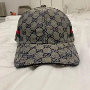 Säljer en snygg Gucci keps med det klassiska GG-monogrammet i blått på beige bakgrund. Kepsen har de ikoniska gröna och röda ränderna på sidan. Perfekt för att ge din outfit en lyxig touch. Justerbar passform för att passa de flesta huvudstorlekar. Självklart är kepsen äkta men tyvärr inget kvitto.