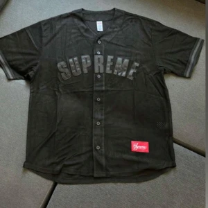 Svart Supreme tröja - Säljer en svart Supreme tröja i mesh-material med knappar framtill. Den har korta ärmar och Supreme-loggan i stort tryck över bröstet. Perfekt för en avslappnad streetwear-look. Tröjan är i mycket bra skick och kommer i originalförpackning.