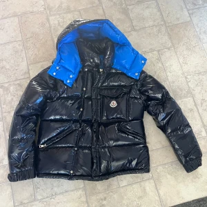 Moncler jacka - Inte äkta. Dustybag o väska följer med!!!!! Säljer en snygg och varm pufferjacka från Moncler i svart med blå insida. Jackan har avtagbara ärmar, vilket gör den superpraktisk för olika väder. Den har en huva och flera fickor med dragkedja. Perfekt för vintern och hösten! 🖤💙