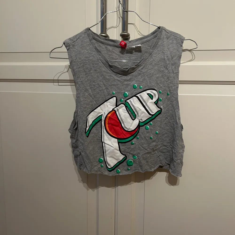 Säljer en cool grå croppad linne med stort 7UP-tryck på framsidan. Perfekt för sommarens varma dagar eller en avslappnad look. Linnet är ärmlöst och har en rå kant nertill för en edgy stil. Passar perfekt till jeans eller shorts!. Toppar.