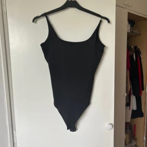Svart linne bodysuit - Snygg svart bodysuit i ett stretchigt material. Den är ärmlös med smala axelband och har en enkel, stilren design. Perfekt för att matcha med jeans eller kjol för en chic look. Passar både till vardags och fest!
