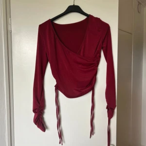 Vinröd omlottopp med knytband - Snygg vinröd omlottopp i mjukt material med långa ärmar. Toppen har knytband vid ärmsluten och en smickrande omlottdesign framtill. Perfekt för höst och vinter, både till vardags och fest. Passar bra med jeans eller kjol för en chic look!