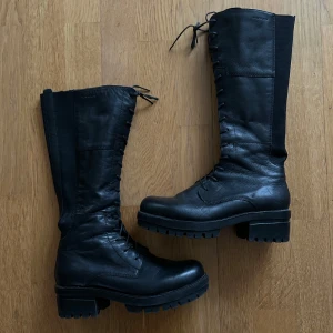 Svarta höga boots med snörning - Säljer ett par svarta höga boots i skinn med snörning och dragkedja på sidan. De har en robust sula och är perfekta för höst och vinter. Insidan har ett snyggt rutigt mönster. Skorna är i mycket bra skick och redo för nya äventyr!