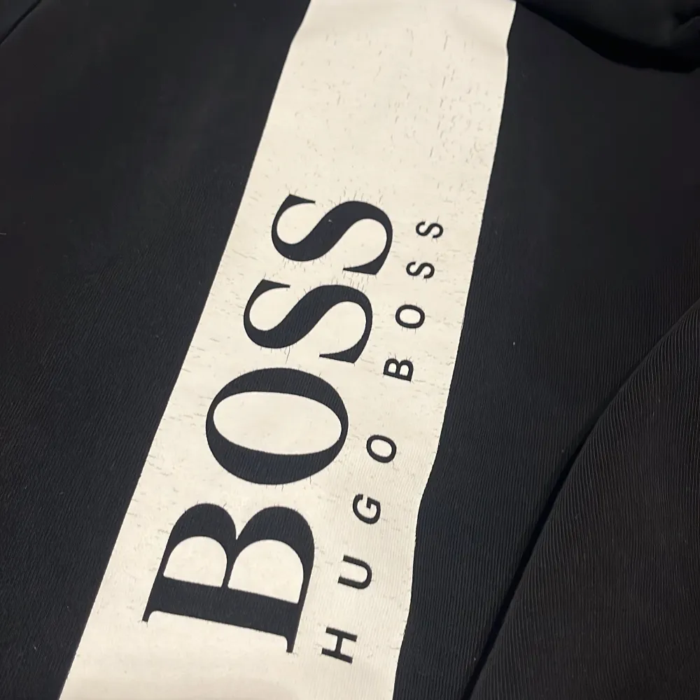 Säljer en stilren svart hoodie från Hugo Boss i mycket bra skick. Den har en dragkedja framtill och en stor logga på ryggen. Perfekt för höst och vinter, med långa ärmar och en bekväm passform. Huvan har vita snören som en snygg detalj. Har använt den ganska mycket köpt för 800kr . Tröjor & Koftor.