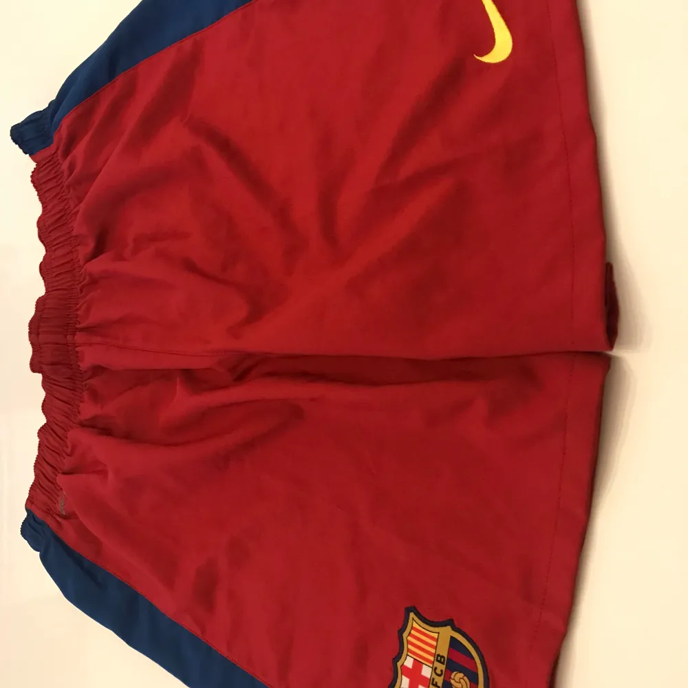 Säljer ett par röda Barcelona shorts som är köpta på camp now. De har en elastisk midja och är perfekta för fotbollsträning. Shortsen har blå detaljer på sidorna och klubbens emblem på ena benet. De är i bra skick och redo för nya äventyr på planen! ⚽️. Shorts.