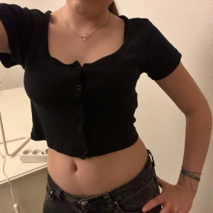 Svart croppad topp med knappar - Säljer en svart croppad topp med knappar framtill. Den har korta ärmar och en v-ringad design. Perfekt för sommardagar eller en avslappnad kvällslook. Toppen är i bra skick och passar till både jeans och kjol.