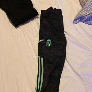 Säljer ett par svarta träningsbyxor från Adidas med Real Madrid-logga. Byxorna har gröna detaljer och klassiska Adidas-ränder längs sidorna. Perfekta för fotbollsträning eller chill. De är i bra skick och har en bekväm passform.