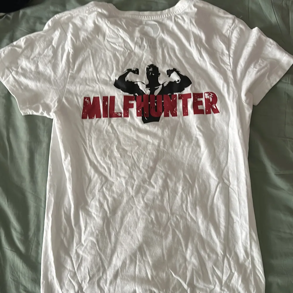 Säljer en vit t-shirt från Revenite i storlek S. Den har ett tryck på baksidan med texten 'MILFHUNTER' . Perfekt för en avslappnad stil eller som statement-plagg. T-shirten är i bomull och har korta ärmar. T-shirts.