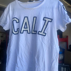 Vit t-shirt med 'CALI'-tryck - Säljer en vit t-shirt från 157 med stort 'CALI'-tryck på framsidan. Den är kortärmad och gjord i bomull, perfekt för en avslappnad stil. T-shirten har en liten fläck på framsidan, men är annars i bra skick. Perfekt för sommardagar eller som en del av en casual outfit.