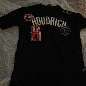 Två coola t-shirts från Hoodrich. Den ena är svart med stora vita och röda bokstäver och flera märken på framsidan. Den andra är ljusblå med texten 'Hoodrich' i svart och grått på framsidan. Perfekta för en avslappnad stil!