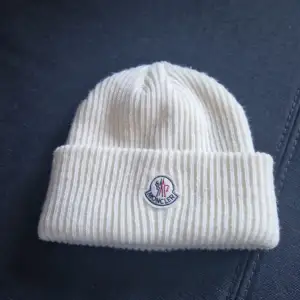 Säljer en superfin vit stickad mössa från Moncler. Den är riktigt mjuk och varm, perfekt för kyliga dagar. Mössan har Monclers klassiska logga broderad på framsidan. Den är i nyskick och känns lyxig att ha på sig. Passar perfekt till både vardag och lite finare tillfällen.