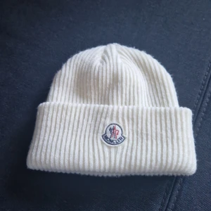 Moncler mössa - Säljer en superfin vit stickad mössa från Moncler. Den är riktigt mjuk och varm, perfekt för kyliga dagar. Mössan har Monclers klassiska logga broderad på framsidan. Den är i nyskick och känns lyxig att ha på sig. Passar perfekt till både vardag och lite finare tillfällen.