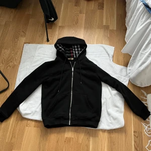 Svart hoodie från Burberry - Säljer en stilren svart hoodie från Burberry med dragkedja och klassiskt rutigt mönster i huvan. Perfekt för en avslappnad och trendig look. Hör av dig vid intresse! Storlek L men sitter som s/m