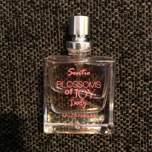 Blossoms of Joy Lovely Eau de Parfum - Elegant parfymflaska med en rektangulär form och en stilren design. Flaskan är genomskinlig med rosa text och en silverfärgad spraykork. Doften heter 'Blossoms of Joy Lovely' och är en Eau de Parfum, vilket ger en långvarig och intensiv doftupplevelse. Har knappt använt den (se bild)😊