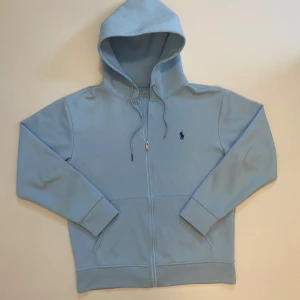 Ljusblå Ralph Lauren zip hoodie - Säljer en stilren ljusblå zip up hoodie från Polo Ralph Lauren med dragkedja och broderad logga på bröstet. Perfekt för en avslappnad stil med ribbade muddar och fickor framtill. Huvan har dragsko för justerbar passform. Använd max tre gånger men saknar en metall bit på snöret men inget som märks när den är på, skriv vid minsta fråga eller fundering💯👍