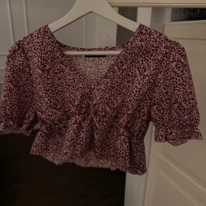 Blommig blus med volangdetaljer - Söt blommig blus i rosa och svart med volangdetaljer. Blusen har en v-ringad design och korta arm