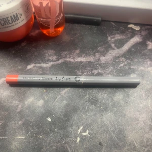 Läpppenna från The Makeup Library - En läppenna i en livfull orange nyans från The Makeup Library. Pennan har en grå hylsa med en tydlig märkning och en skruvbar funktion för enkel applicering. Någon slags orange/rosa färg på läppenan!🫶😊❤️