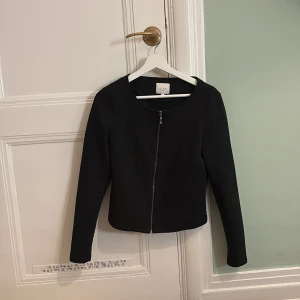 Svart hoodie med dragkedja från Vila - Snygg svart hoodie från Vila med dragkedja framtill. Jackan har en quiltad textur och långa ärmar, perfekt för en stilren look. Passar bra till både vardag och mer uppklädda tillfällen.