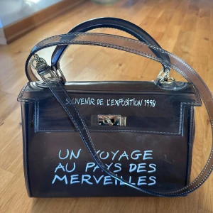 Transparent handväska med text - Snygg transparent handväska med texten 'Un voyage au pays des merveilles' och 'Souvenir de l'exposition 1990' i vitt. Väskan har ett elegant handtag och en axelrem i grått syntetmaterial. Den har en metallåsdetalj framtill.
