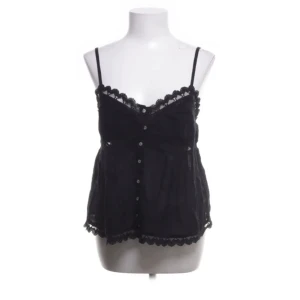 Svart spetslinne med knappar - Snyggt svart linne med smala axelband och dekorativa spetsdetaljer längs kanterna. Linnet har knappar framtill och en luftig passform, perfekt för varma dagar.