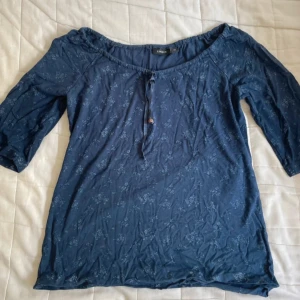Blå blommig topp från Lindex - Säljer en blå topp från Lindex med ett diskret blommönster. Toppen har trekvartsärm och en liten knytning framtill vid halsringningen. Söt topp som går att ha både off shoulder eller på💕Lite sliten ner till men det är på insidan av tröjan så inget man ser!