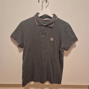 Grå pikétröja från Moncler - Snygg grå pikétröja från Moncler med klassisk krage och knappar. Tröjan har en broderad logotyp på bröstet och röd-vit randdetalj på krage och ärmslut. Perfekt för en stilren look.