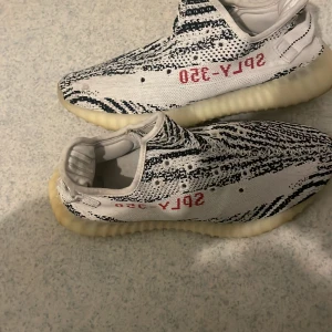 Adidas Yeezy Boost 350 V2 Zebra - Säljer ett par Adidas Yeezy Boost 350 V2 i Zebra-mönster. Skorna har en vit och svart design med röd text på sidan. De är tillverkade i syntetmaterial och har en bekväm, ribbad sula. Storlek 42/41.5. Pris kan diskuteras. Förekommer kvitto!