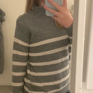 Grå och vit randig tröja med guldiga knappar - Säljer en grå tröja med vita ränder och dekorativa knappar på axeln. Tröjan har även en hög krage. Det står tyvärr inte vart den är ifrån.  Fråga ifall ni undrar nått mått🤍🤍