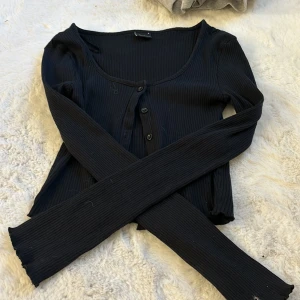 Svart ribbad kofta från Gina Tricot - Säljer en stilren svart ribbad kofta från Gina Tricot. Koftan har långa ärmar och knappar framtill, perfekt för lager-på-lager-stil. Ett mångsidigt plagg för din garderob!