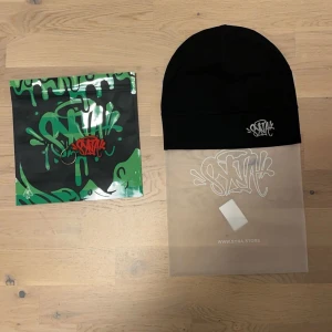 Svart mössa från SYNA ( Central Cees märke) - Snygg svart mössa från SYNA med broderad logga framtill. Tillverkad i en mjuk blandning av polyester och polyamid för en bekväm passform. Perfekt för att hålla värmen med stil.