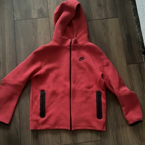 Röd hoodie från Nike - Snygg röd hoodie från Nike tech fleece med dragkedja och huva. Jackan har svarta detaljer vid fickorna och Nike-logga på bröstet. Perfekt för en sportig look eller avslappnad stil. I superfint skick. Inga fläckar eller noppor.