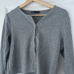 Grå ribbad kofta från Zara - Säljer en stilren grå ribbad kofta från Zara. Den har en croppad passform med knappar framtill och långa ärmar. Perfekt för lager-på-lager-stil eller som ett lättare ytterplagg.