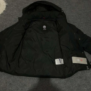  Canada goose wyndham parka  - Säljer en svart dunjacka från Canada Goose wynd ham parka med luva och broderad logotyp på ärmen. Jackan har långa ärmar och stängs med dragkedja och knappar. Perfekt för kalla dagar. 