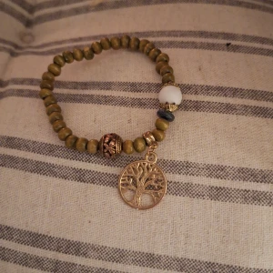 Armband med livets träd-berlock - Unikt armband med gröna pärlor och en vacker livets träd-berlock. Armbandet har även en stor vit pärla och en dekorativ metallpärla. Perfekt för att ge en naturlig touch till din stil.