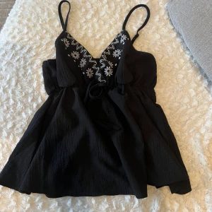 Svart linne med broderi - Snyggt svart linne med smala axelband och v-ringning. Detaljerat med vackert vitt broderi längs halsringningen. Perfekt för varma dagar eller som ett lager under en jacka.