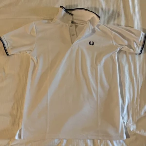 Vit pikétröja från Fred Perry - Snygg vit pikétröja från Fred Perry med klassisk krage och knappar. Tröjan har en broderad logotyp i marinblått på bröstet och kontrasterande ränder på kragen och ärmsluten. Om du undrar något är det bara att höra av sig, har även fler storlekar i lagret!