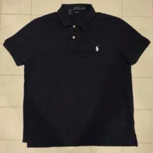 Svart pikétröja från Ralph Lauren - Snygg Mörkblå pikétröja från Ralph Lauren med klassisk passform. Tröjan har korta ärmar och en broderad logotyp i vitt på bröstet. Perfekt för en stilren look.