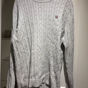 Grå kabelstickad tröja från Bondelid - Säljer en stilren grå kabelstickad tröja från Bondelid. Tröjan har en rund halsringning och ett litet emblem på bröstet. Perfekt för kyligare dagar.