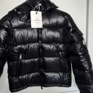 Moncler Maya - Snygg svart pufferjacka från Moncler med glansig finish. Jackan har en dragkedja framtill och praktiska fickor med dragkedjor. Perfekt för kyliga dagar med sin vadderade design.               (Storlek: M L XL 2XL