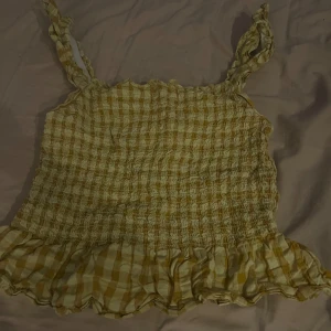 Gul rutig topp med volang - Charmig gul rutig topp med volangdetalj i nederkant. Toppen har smala axelband och en luftig passform, perfekt för varma sommardagar.