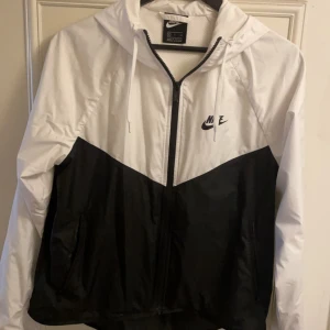 Svartvit vindjacka från Nike - Snygg svartvit vindjacka från Nike med dragkedja och huva. Jackan har en klassisk design med Nike-logga på bröstet och praktiska fickor med dragkedja. Perfekt för blåsiga dagar. Använd typ 1 vecka men passade inte så bra till vintern för mig