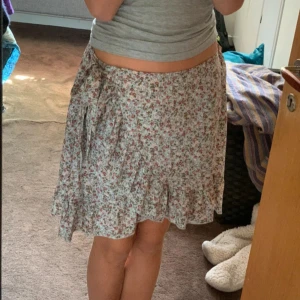 Blommig kjol med volang - Söt blommig kjol i ljusa färger med volangdetaljer. Kjolen har en omlott-design och knytning i sidan, vilket ger en romantisk känsla. Perfekt för varma dagar.