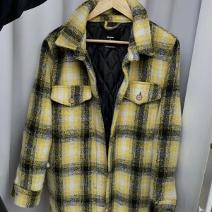 Rutig jacka från bikbok - Snygg rutig jacka från Bikbok i gula och svarta toner. Jackan har en quiltad insida och knappar framtill. Perfekt för höst och vår med sin stilrena design och bekväma passform.