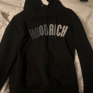 Svart hoodie från Hoodrich - Säljer en svart hoodie från Hoodrich med stor logga på bröstet. Den har en klassisk känguruficka och justerbar huva med dragsko. Perfekt för en avslappnad stil.